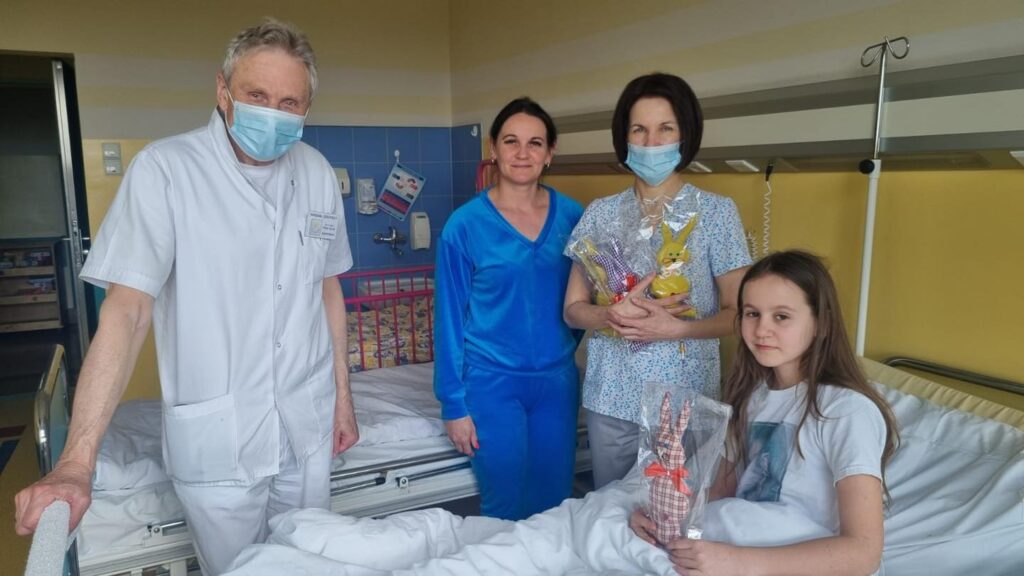 Dziecko w szpitalnym łóżku z zabawką-zającem. Obok mama, pielęgniarka i lekarz.